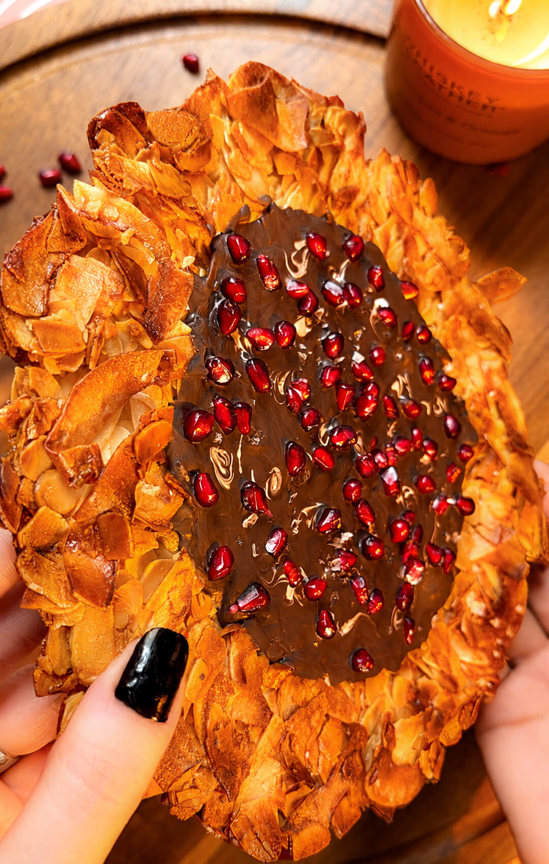 A close-up shot of hands holding up a whole crunchy Florentine cookie pizza wheel to show its crisp, lacy, golden texture. Made with sliced almonds, coconut flakes, a rich dark chocolate centre, and a sprinkling of fresh pomegranate seeds.