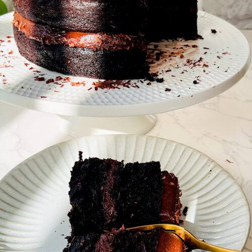 A slice of healthy moist chocolate cake with a gold fork next to a whole two layer paleo gluten free cake made with almond flour and coconut flour covered in silky dairy free chocolate ganache.