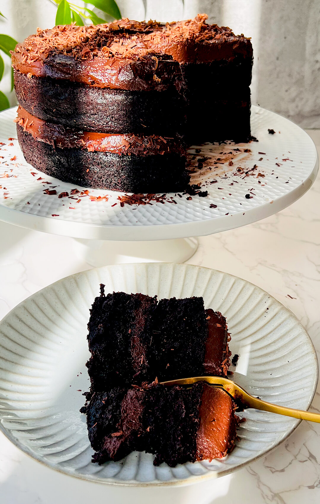 A slice of healthy moist chocolate cake with a gold fork next to a whole two layer paleo gluten free cake made with almond flour and coconut flour covered in silky dairy free chocolate ganache.
