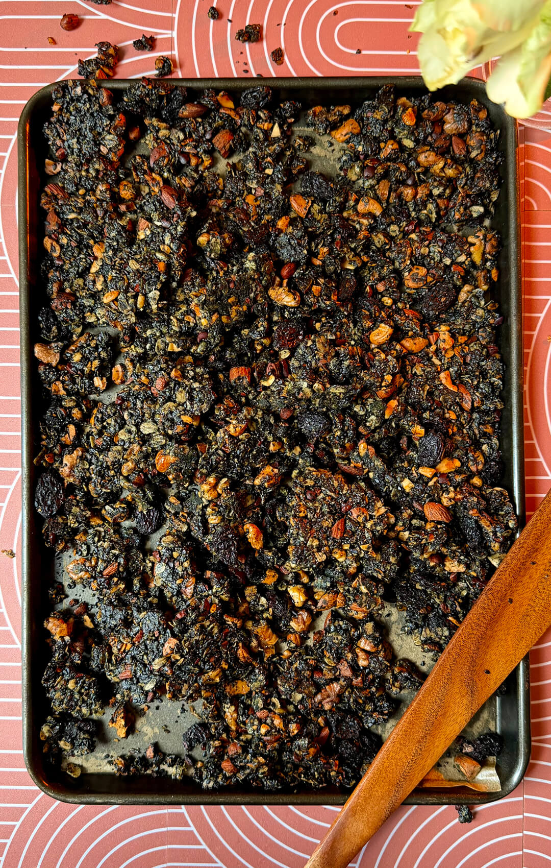: A hero image of crispy black tahini granola clusters spread across a baking tray on a patterned backdrop beside a wooden spoon. This homemade superfood granola is rich in healthy fats, plant-based protein, fiber, and omega-3 fats from walnuts. Made with rolled oats, black tahini, black sesame seeds, poppy seeds, roasted almonds, cashews, sunflower seeds, black raisins, cacao nibs, honey, and coconut oil, this recipe makes a nutritious gluten-free breakfast or healthy snack.