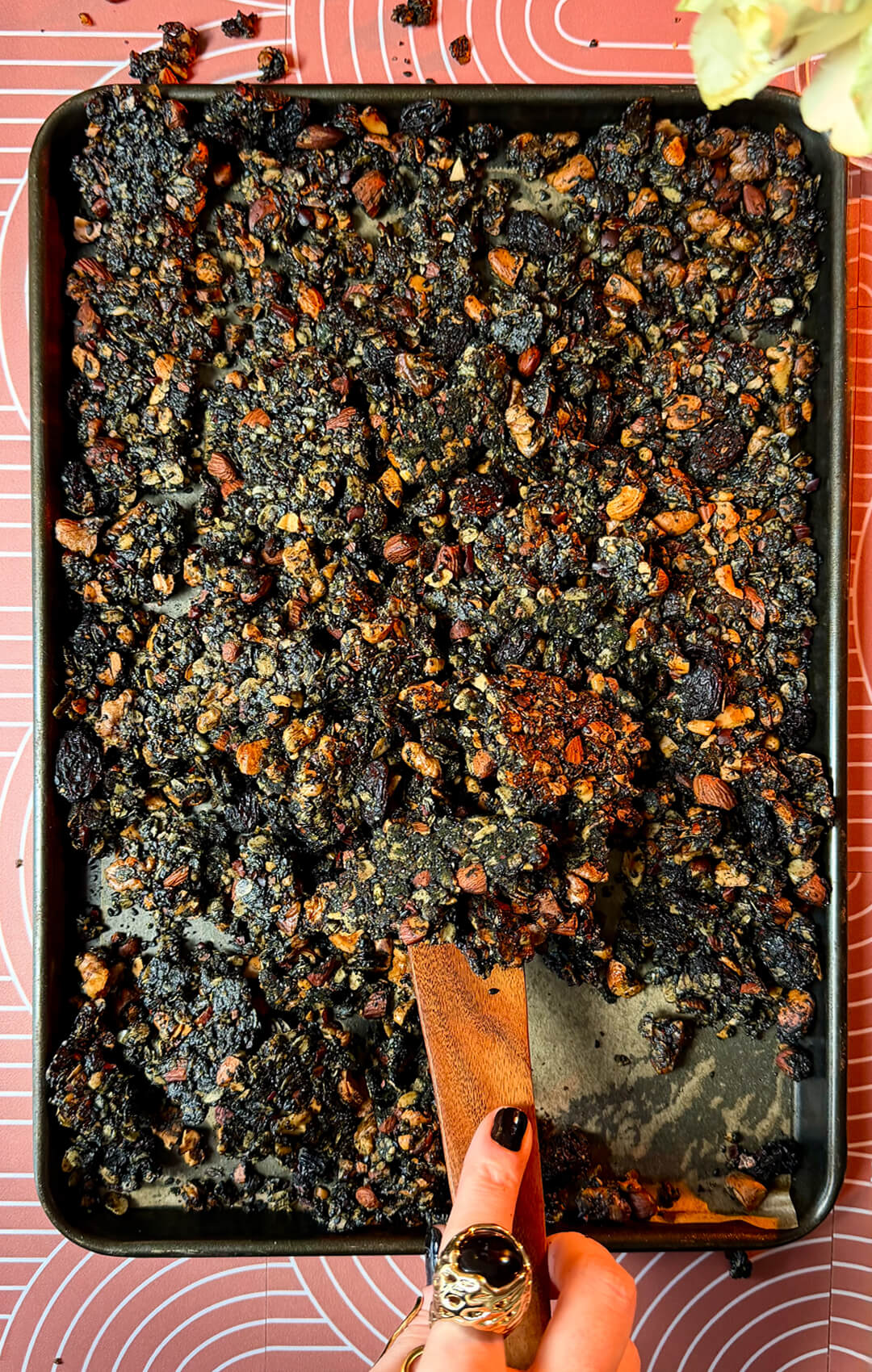 : Freshly baked sheet pan of crunchy, crispy black tahini granola clusters being scooped with a wooden spatula. This wholesome homemade superfood granola recipe features gluten-free oats, roasted almonds, walnuts, cashews, sunflower seeds, cacao nibs, black sesame seeds, poppy seeds, black raisins, black tahini, coconut oil, natural honey, and warm spices for a nutritious gluten-free and refined sugar-free breakfast or healthy snack.