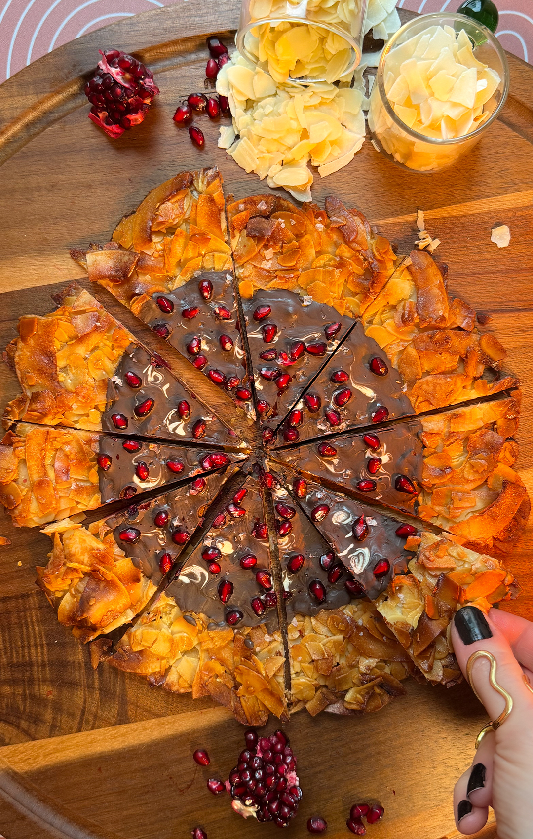 An overhead shot of a hand with black nail polish pulling a single wedge from a sliced crispy Florentine cookie pizza. Flaky toasted almonds, crispy coconut flakes, and glints of melted dark chocolate show off the easy-to-share dessert style. this healthy cookie wheel is paleo, gluten-free, refined sugar free. quick and easy to make at home with 7 simple ingredients and great for meal prep.