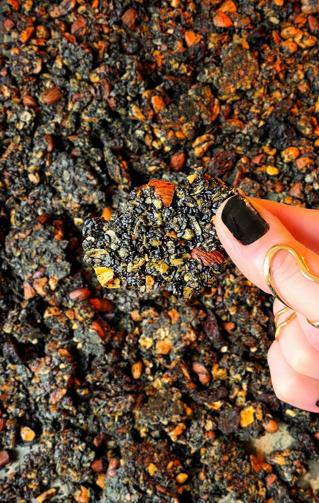 A close-up shot of a hand holding a large chunky black tahini granola cluster over a baking tray. This ultra-crunchy homemade granola is naturally sweetened with honey, baked with coconut oil, and packed with roasted cashews, walnuts, almonds, sunflower seeds, cacao nibs, black sesame seeds, poppy seeds, black tahini, black raisins, and warming spices, making it a wholesome gluten-free breakfast, healthy snack, or nutrient-dense superfood treat.