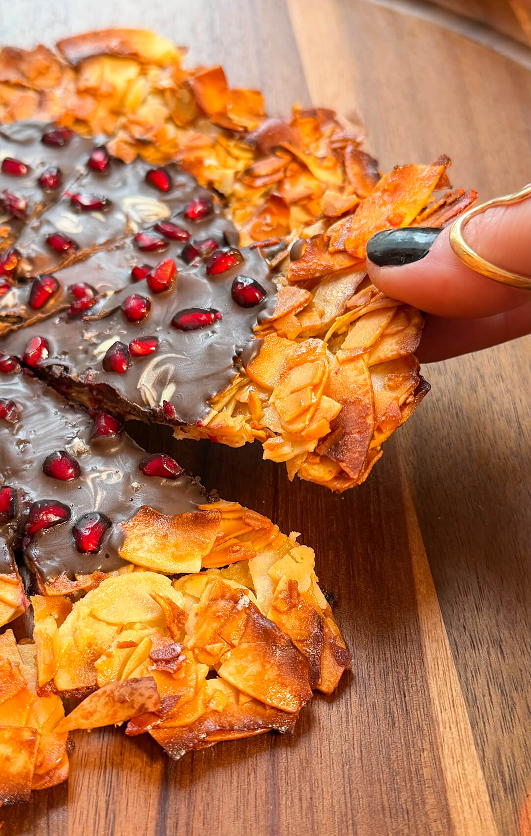 A close-up side angle of a hand lifting a thin, crispy Florentine cookie pizza wedge from a wooden platter. The shot highlights the delicate, crispy and lacy texture of the toasted almonds and the solid layer of dark chocolate.so easy to make at home with just 7 simple ingredients. this is healthy, gluten-free, paleo and refined sugar free.