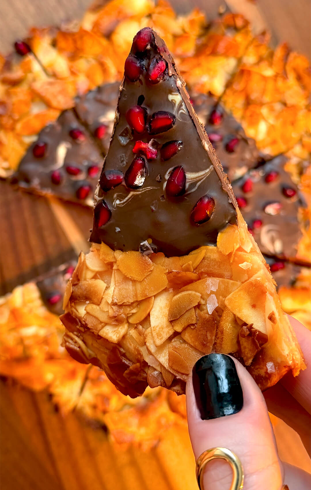 A hand holding a single triangular slice of crunchy Florentine cookie pizza up to the camera. The slice features a thick coating of dark chocolate packed with ruby-red pomegranate arils (seeds) and large flakes of sea salt over a base of sliced almonds.