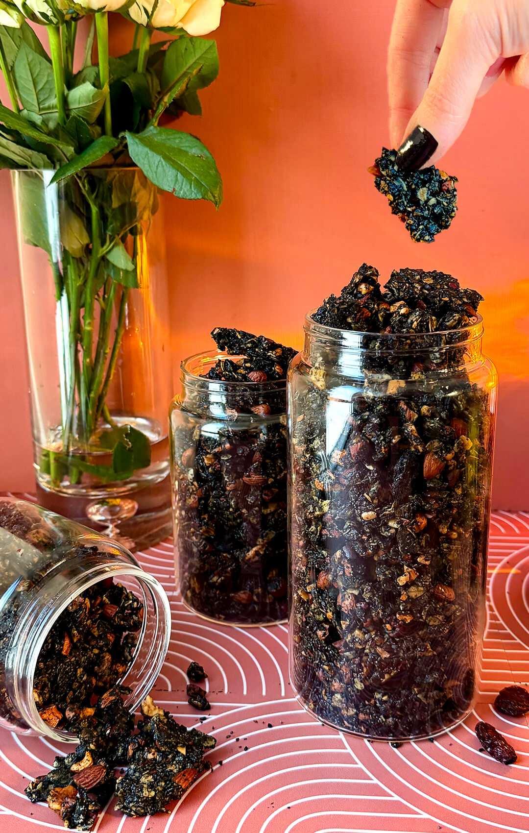 A hand with black nail polish picking a crunchy black tahini granola cluster from a tall glass storage jar filled with homemade granola. This wholesome superfood snack is made with rolled oats, roasted nuts, sunflower seeds, black sesame seeds, poppy seeds, black raisins, cacao nibs, black tahini, coconut oil, honey, and warming spices for a gluten-free breakfast, healthy snack, or refined sugar-free treat.