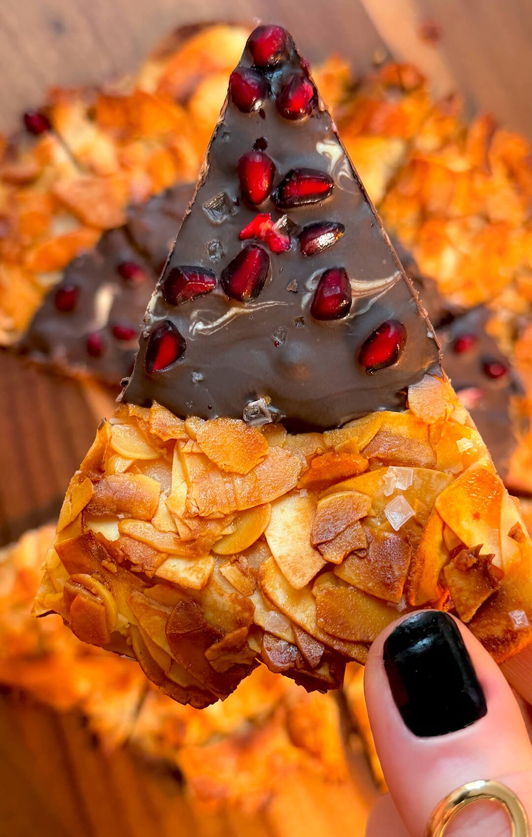 A detailed close-up shot of a paleo Florentine cookie pizza wedge held in focus. The view showcases the golden-brown caramelized almond and coconut crust topped with glossy dark chocolate, bright pomegranate seeds, and coarse sea salt flakes.