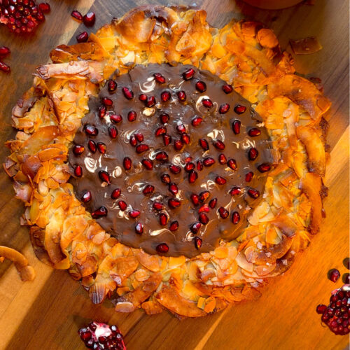 Quick and easy crunchy Florentine cookie pizza made at home with toasted almond slices and coconut flakes, featuring a golden crisp texture with a soft chewy centre, coated in dark chocolate and fresh pomegranate arils (seeds). This healthy recipe is gluten-free, paleo, and refined sugar-free.