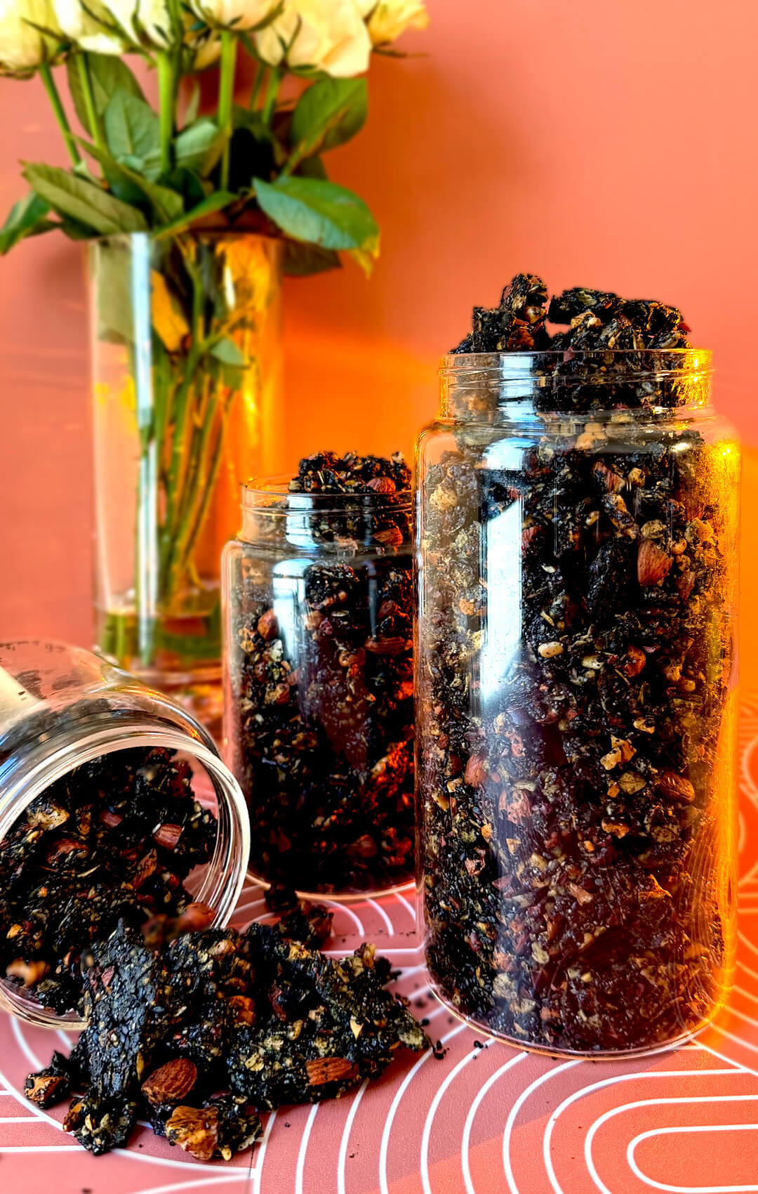 Two upright glass storage jars beside an overturned jar spilling crunchy black tahini granola clusters onto a patterned surface next to white roses. This nutrient-dense homemade granola features rolled oats, roasted almonds, cashews, walnuts, sunflower seeds, black sesame seeds, poppy seeds, black raisins, cacao nibs, black tahini, coconut oil, and honey for a wholesome gluten-free breakfast, healthy snack, or meal-prep treat.