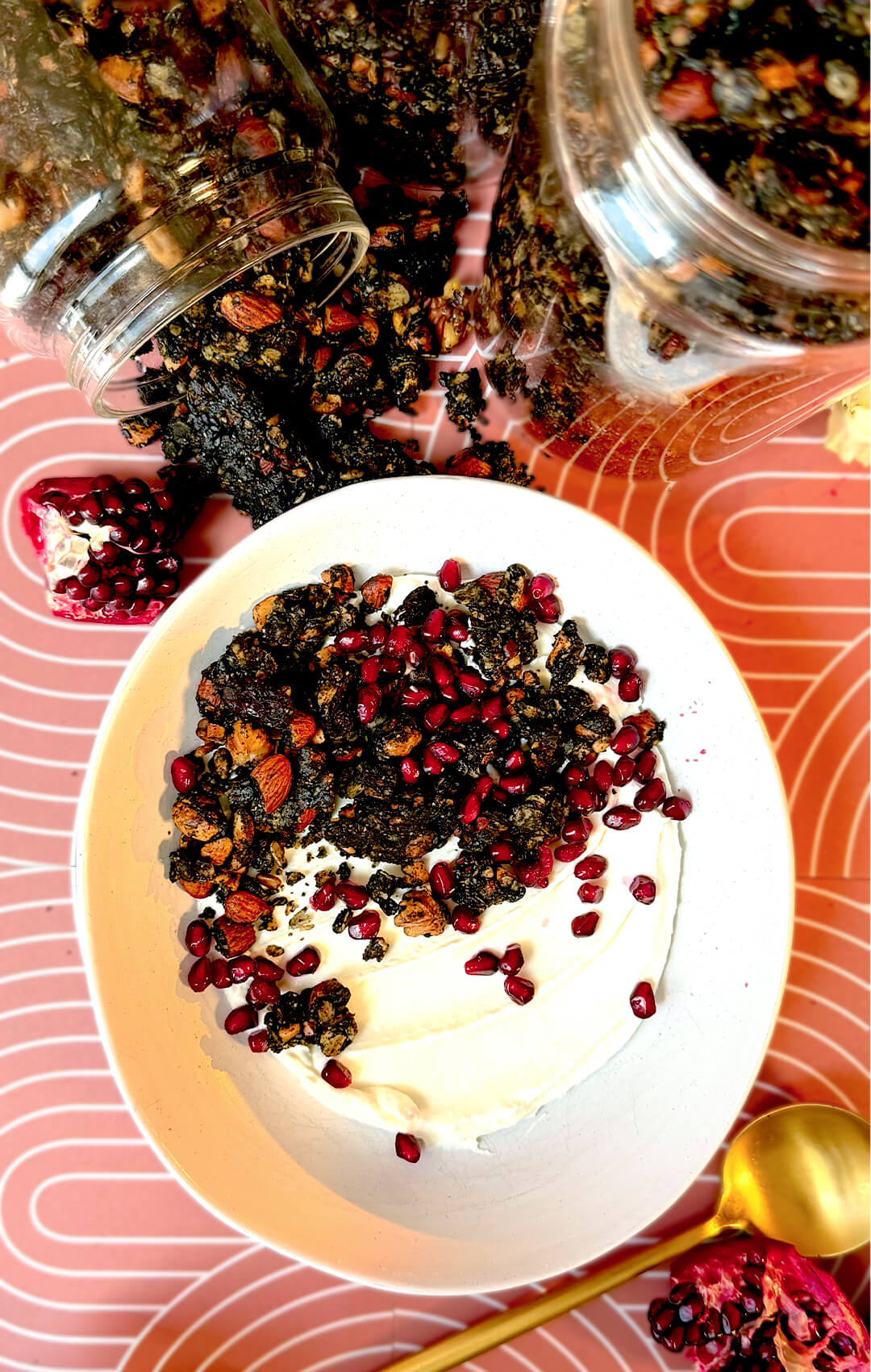 A top-down view of a bowl of creamy yogurt topped with crunchy black tahini granola clusters and fresh pomegranate seeds. This high-fiber breakfast and plant-based protein snack features black tahini, black sesame seeds, poppy seeds, rolled oats, roasted almonds, walnuts, cashews, black raisins, and cacao nibs for sustained energy, digestive support, and a nutritious gluten-free meal.
