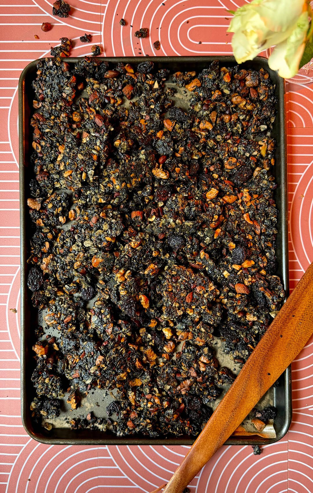A hero image of crispy black tahini granola clusters spread across a baking tray on a patterned backdrop beside a wooden spoon. This homemade superfood granola is rich in healthy fats, plant-based protein, fiber, and omega-3 fats from walnuts. Made with rolled oats, black tahini, black sesame seeds, poppy seeds, roasted almonds, cashews, sunflower seeds, black raisins, cacao nibs, honey, and coconut oil, this recipe makes a nutritious gluten-free breakfast or healthy snack.