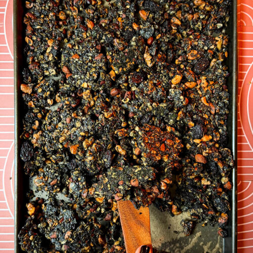 Freshly baked sheet pan of crunchy, crispy black tahini granola clusters being scooped with a wooden spatula. This wholesome homemade superfood granola recipe features gluten-free oats, roasted almonds, walnuts, cashews, sunflower seeds, cacao nibs, black sesame seeds, poppy seeds, black raisins, black tahini, coconut oil, natural honey, and warm spices for a nutritious gluten-free and refined sugar-free breakfast or healthy snack.