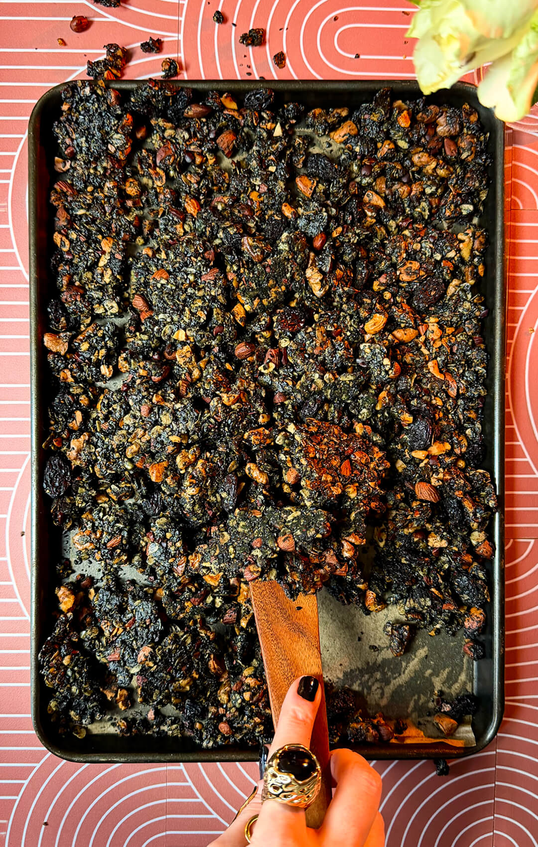 Freshly baked sheet pan of crunchy, crispy black tahini granola clusters being scooped with a wooden spatula. This wholesome homemade superfood granola recipe features gluten-free oats, roasted almonds, walnuts, cashews, sunflower seeds, cacao nibs, black sesame seeds, poppy seeds, black raisins, black tahini, coconut oil, natural honey, and warm spices for a nutritious gluten-free and refined sugar-free breakfast or healthy snack.