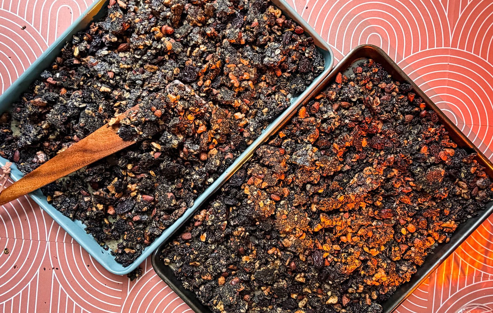 A high-angle, close-up photograph showcasing crispy big cluster granola cooling on two dark baking trays over a pink patterned background. A rustic wooden spoon gently breaks apart the dense, dark purple-black shards. The chunky clusters are tightly packed with healthy ingredients like gluten-free rolled oats, roasted cashews, whole almonds, walnuts, high-fiber sunflower seeds, black sesame seeds, poppy seeds, chewy black raisins, and cocoa powder. This refined sugar-free, superfood granola looks exactly like rustic chocolate bark and is cooling completely to ensure a maximum crunchy, chunky texture for breakfast cereal or yogurt bowls.