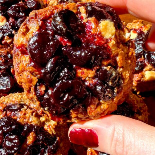 Close up of a healthy peanut butter sour cherry oatmeal cookie with glossy homemade cherry filling, sugar-free white chocolate chips and golden caramelized edges, made with rolled oats and almond flour for a gluten-free, refined sugar-free cookie.