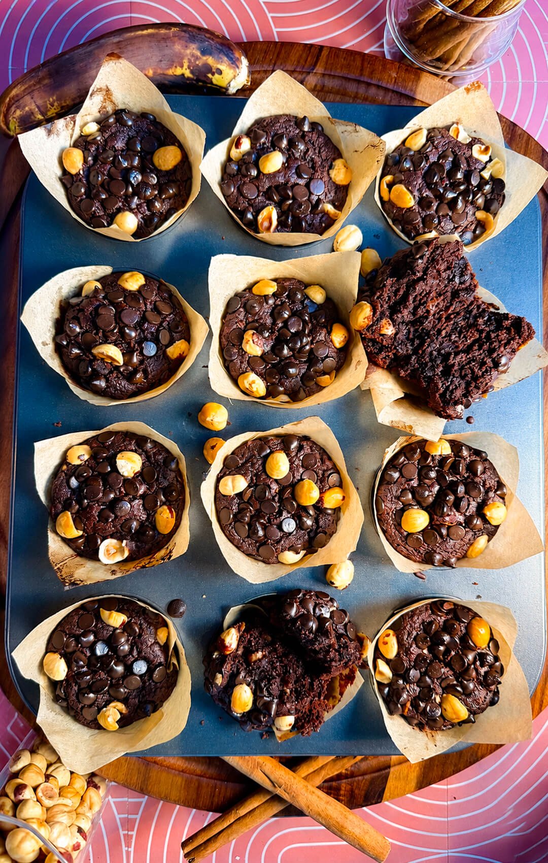 Freshly baked gluten-free double chocolate banana muffins topped with chocolate chips and roasted hazelnuts in parchment liners. one cut to half and show the soft and Fudgy texture.