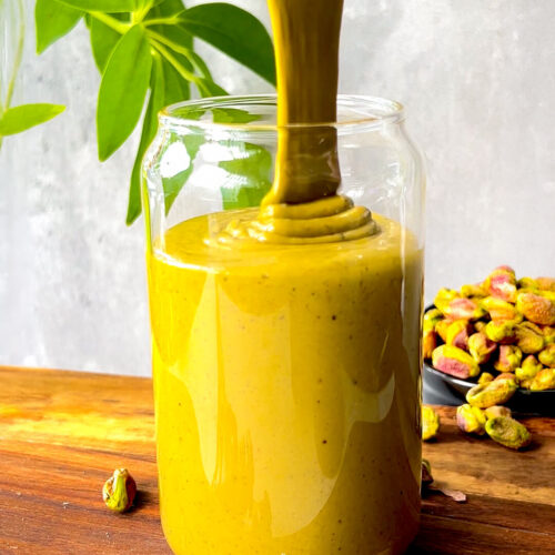 Thick silky homemade pistachio butter pouring into a glass jar, showing its smooth creamy texture, vibrant natural green golden colour, and easy one ingredient pistachio goodness.