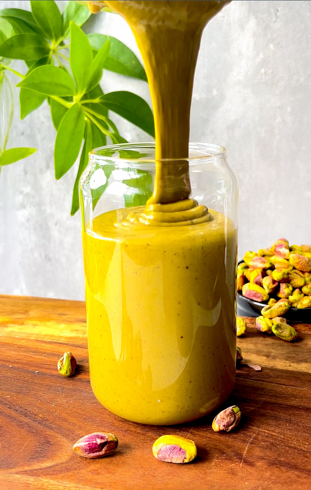Thick silky homemade pistachio butter pouring into a glass jar, showing its smooth creamy texture, vibrant natural green golden colour, and easy one ingredient pistachio goodness.