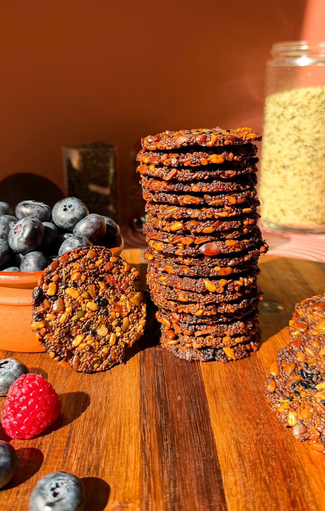 Stack of homemade crispy chia seed crackers with visible pumpkin, sunflower and hemp seeds, served beside fresh blueberries on a wooden board.
