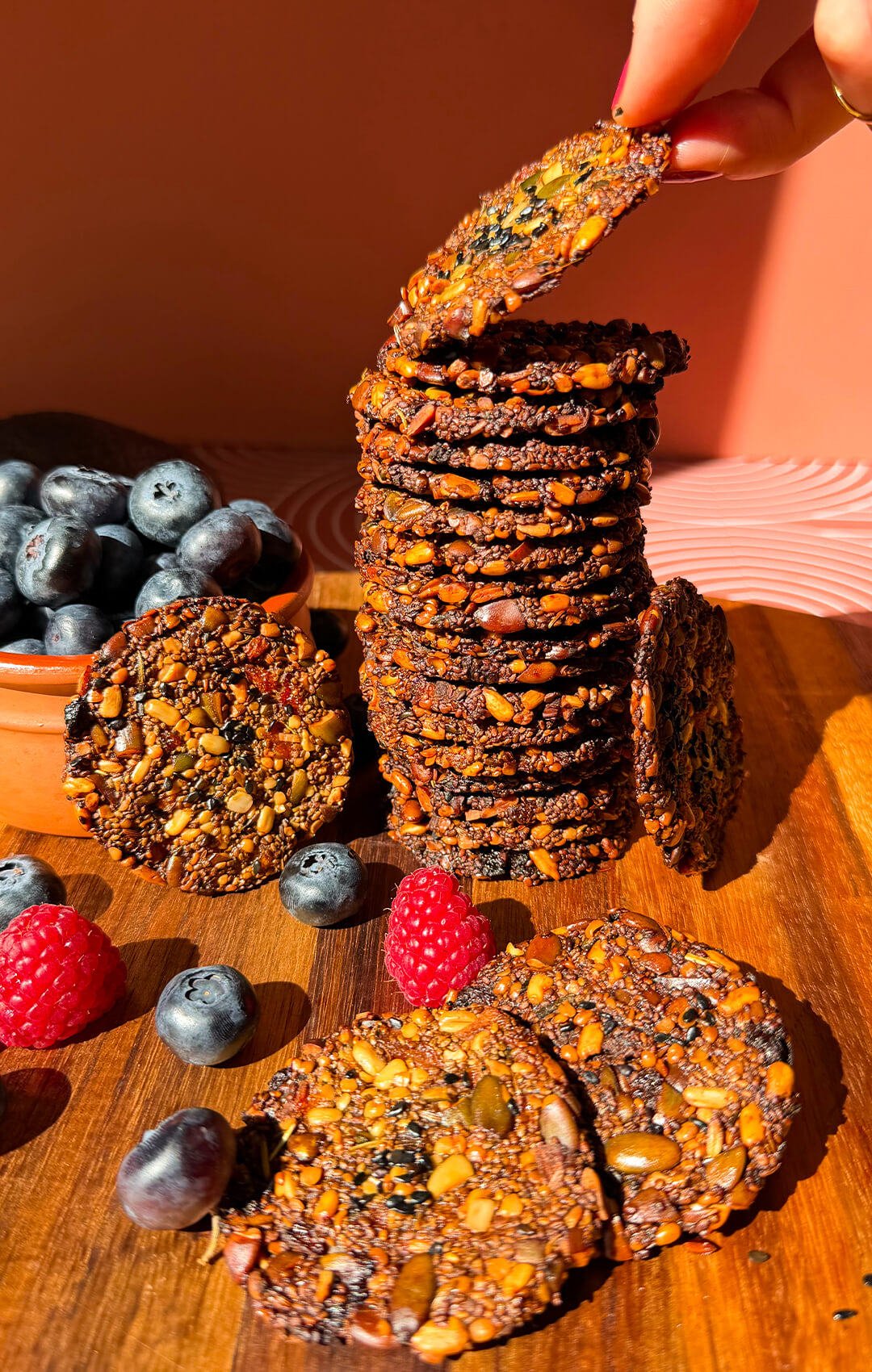 Tall stack of thin crispy chia seed crackers packed with pumpkin, sunflower and hemp seeds, with one cracker being lifted to show its crunchy flourless texture.