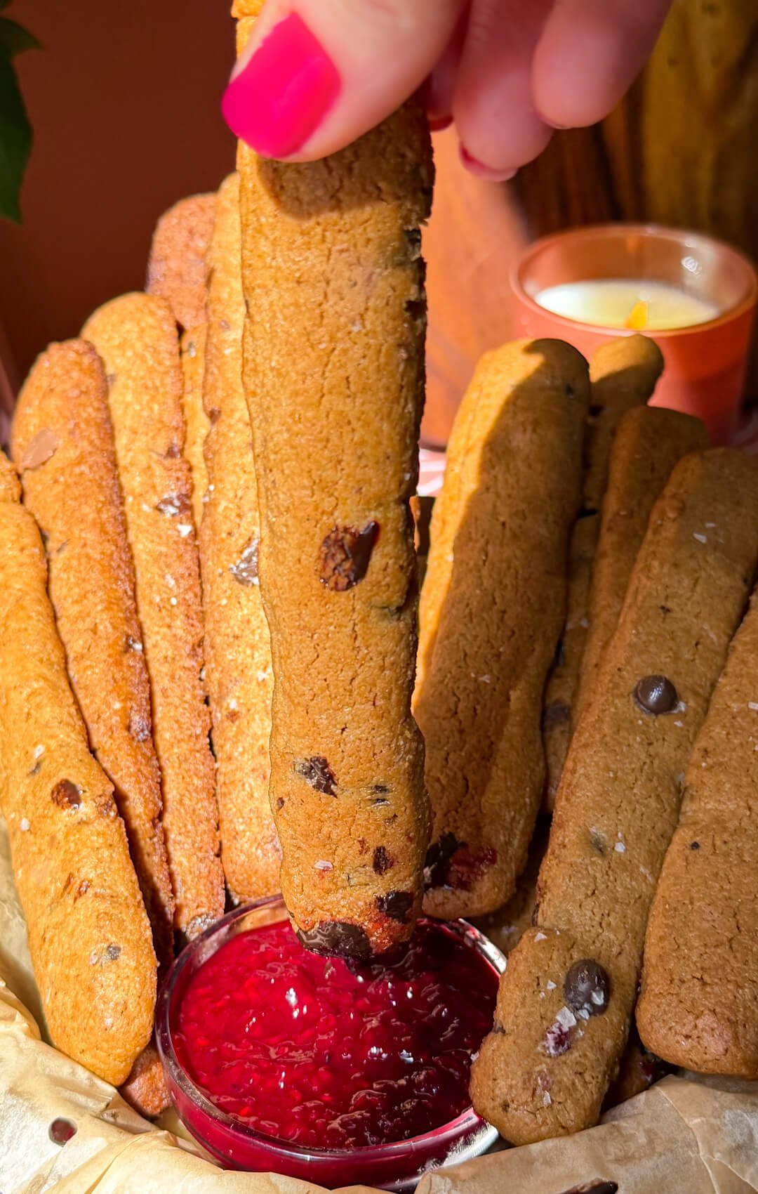 Long golden chocolate chip cookie fry held in one hand, coated at one end with glossy raspberry jam and chocolate sauce, with more cookie fries behind.