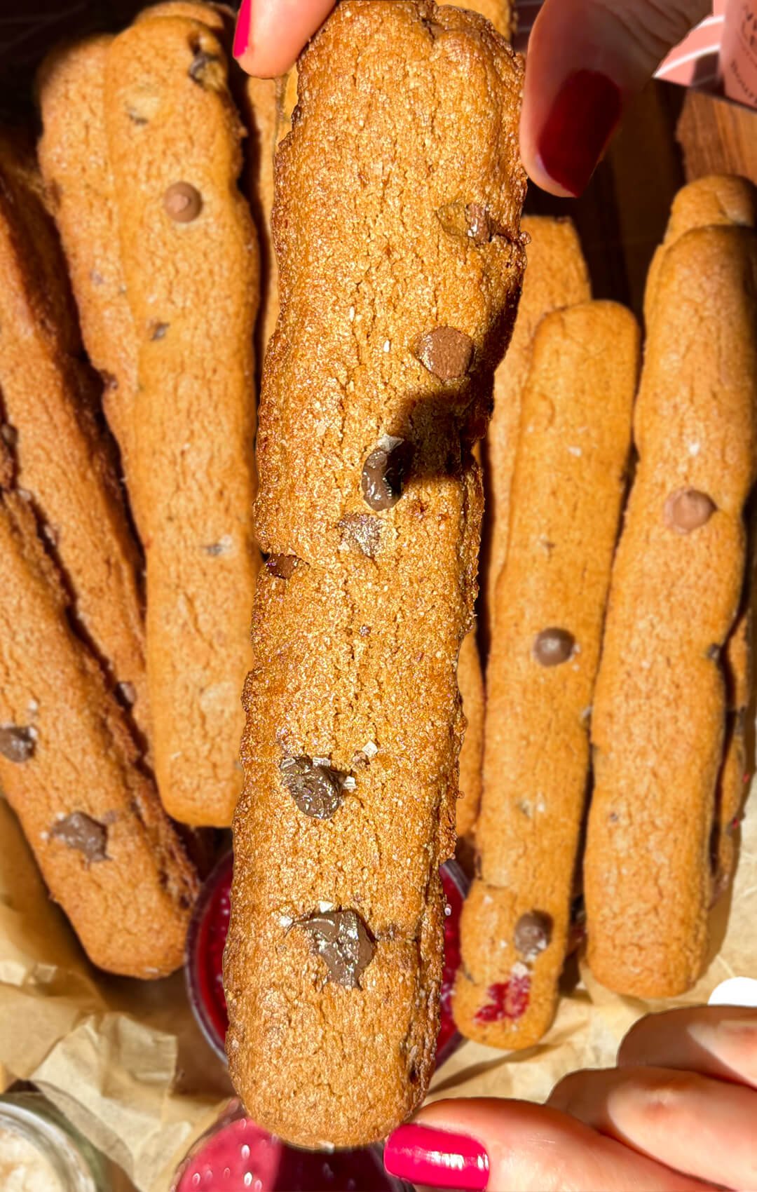 Close-up of a golden brown butter chocolate chip cookie fry held above more gluten-free cookie fries, showing dark chocolate chips and lightly browned edges.