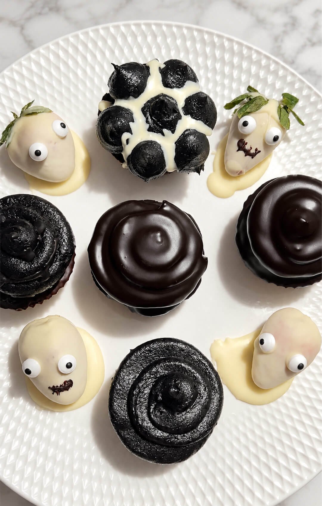 Overhead view of Halloween chocolate cupcakes on a white platter, with shimmering black buttercream swirls, dark chocolate coatings, and white chocolate strawberry ghosts with candy eyes. Easy decorating ideas for gluten-free almond and lupin flour cupcakes with monk fruit chocolate buttercream.
