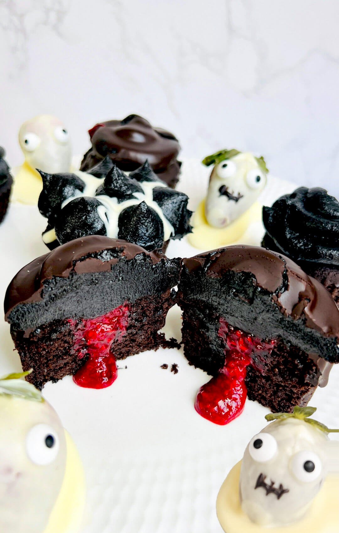 A chocolate-coated Halloween cupcake cut in half reveals a moist chocolate crumb, bright raspberry filling, and a thick layer of black chocolate buttercream. White chocolate strawberry ghosts surround the gluten-free cupcake, made with almond and lupin flours and topped with monk fruit frosting.