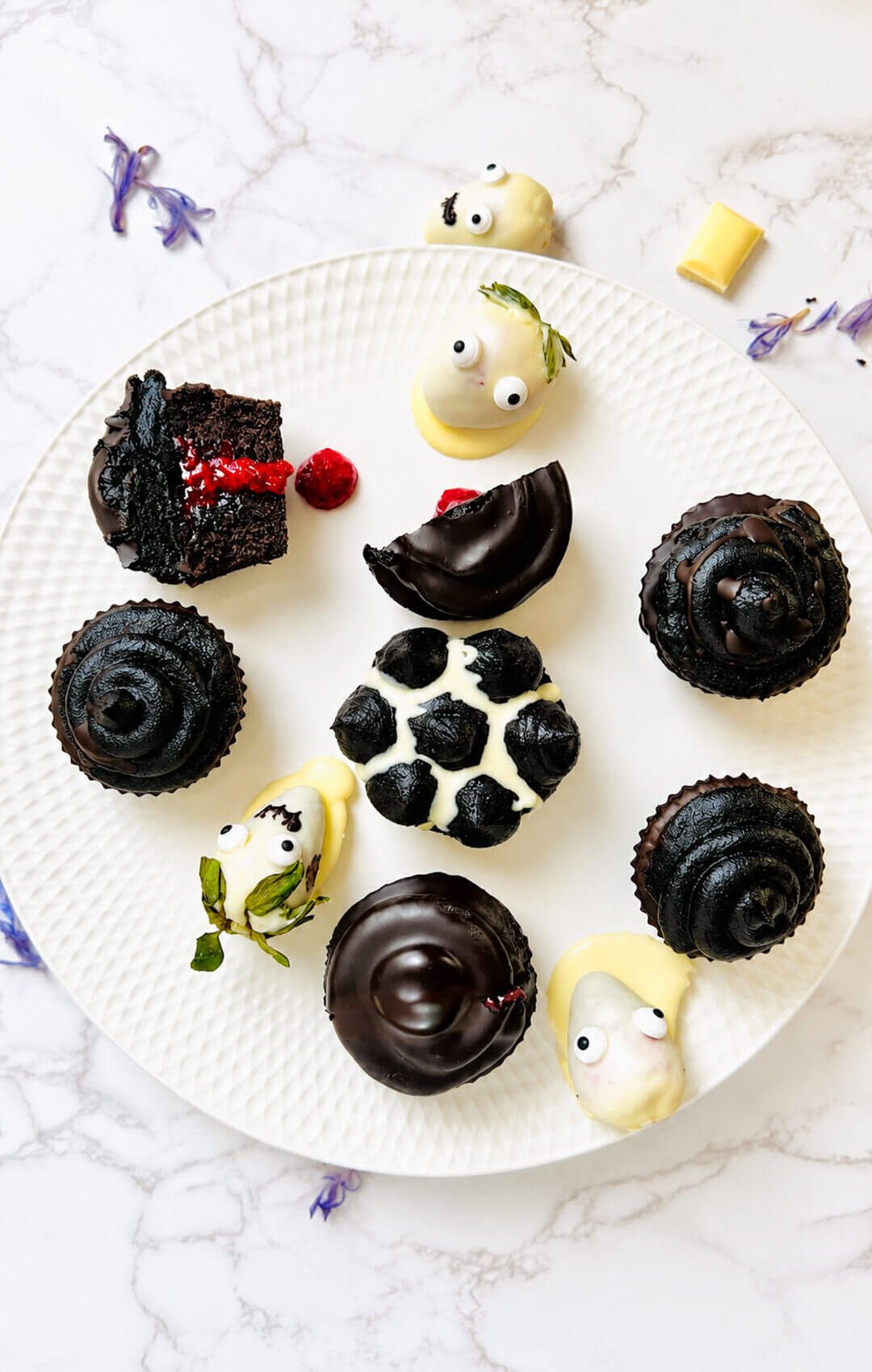 Overhead view of a white platter with black buttercream cupcakes, dark chocolate coatings, white chocolate strawberry ghosts, and a cut cupcake showing its raspberry filling. A mix of simple Halloween cupcake decorating styles with gluten-free chocolate cupcakes and monk fruit frosting.