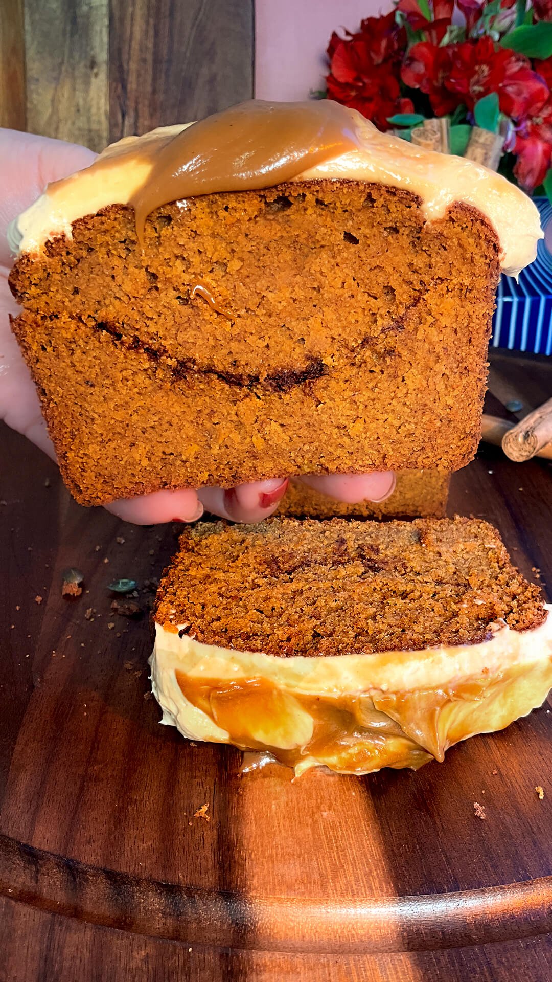 A hand holds a thick slice of healthy pumpkin bread, revealing its moist, tender, bakery-style crumb and cinnamon sugar ribbon beneath peanut butter cream cheese frosting and coconut caramel. An easy gluten-free, refined sugar-free recipe made with almond, oat, and lupin flours, coconut sugar, and brown monk fruit sweetener, with a sugar-free option.