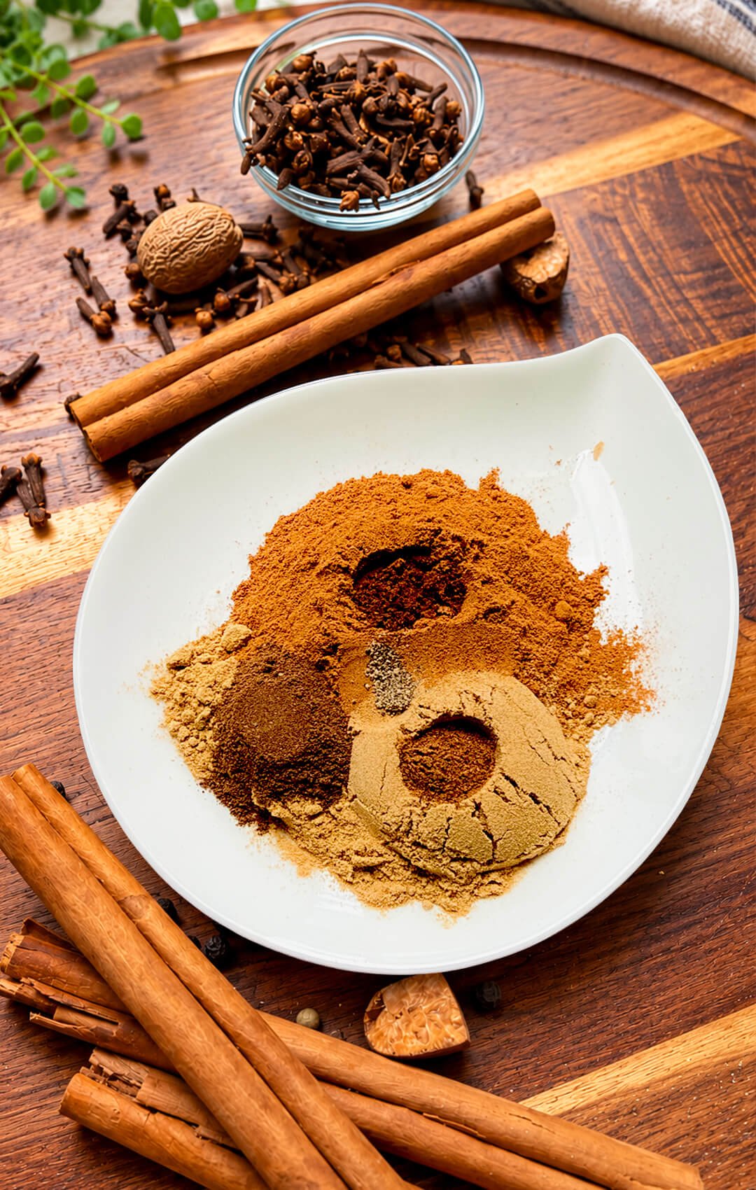 Ground spices in a white bowl with cinnamon sticks, whole nutmeg, and cloves on a wooden board. This easy, aromatic homemade pumpkin pie spice combines cinnamon, ginger, allspice, nutmeg, cloves, and a tiny pinch of black pepper, with no added sugar.