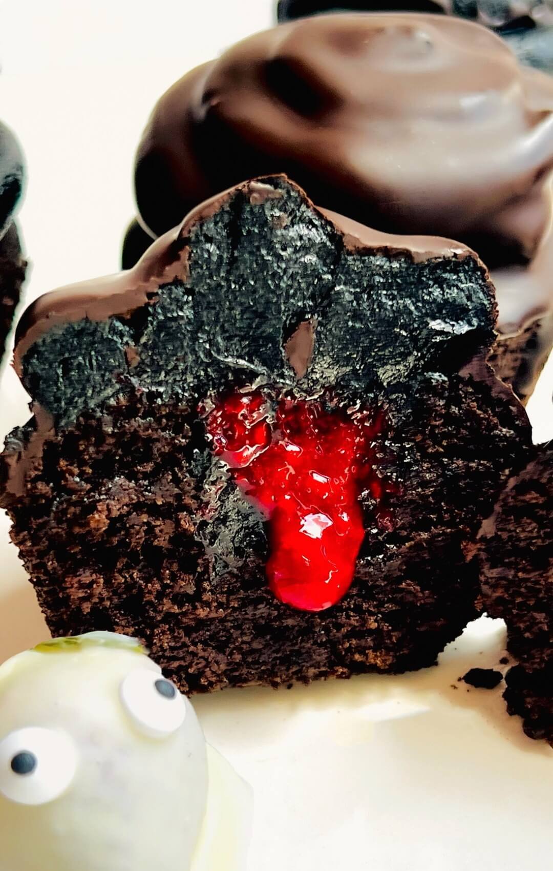 Close-up of a cut Halloween chocolate cupcake showing its moist crumb, red raspberry jam center, thick black chocolate buttercream, and thin dark chocolate shell. A white chocolate strawberry ghost sits beside this gluten-free almond and lupin flour cupcake with sugar-free monk fruit frosting.