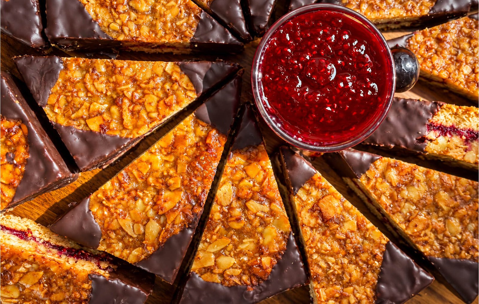 © Healthy Food by Annie. All rights reserved. No reuse, copying, editing, reposting, or redistribution without written permission. A glass bowl of glossy red refined sugar-free raspberry jam surrounded by golden Almond Jam Crunch Cookie Corners with crunchy almond toppings and dark chocolate-dipped tips.