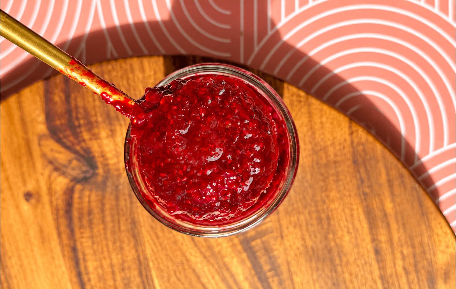 © Healthy Food by Annie. All rights reserved. No reuse, copying, editing, reposting, or redistribution without written permission. Overhead close-up of thick, glossy refined sugar-free raspberry jam with visible fruit pieces and seeds in a glass jar, with a gold spoon resting in the jam.