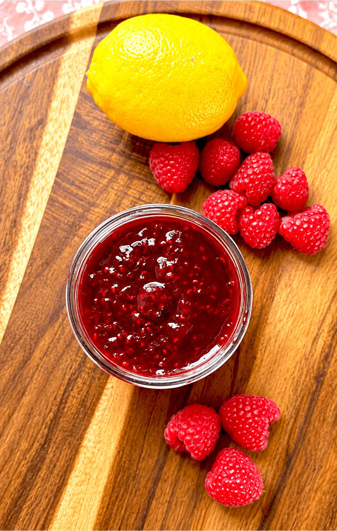 A glass bowl of deep red, glossy refined sugar-free raspberry jam with visible raspberry seeds, surrounded by fresh raspberries and a whole lemon.